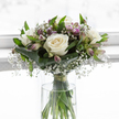 This image captures a softly glowing sympathy bouquet, resting in a clear, cylindrical glass vase that allows the viewer to see the fresh green stems held neatly in clear water. The heart of the arrangement is formed by generous white roses, fully open and velvety, their petals curling outwards in layered spirals that reveal creamy ivory depths and pure white outer edges. Around them, frothy clusters of gypsophila, or baby's breath, add an airy lightness, their countless tiny blossoms giving the bouquet a misty, cloud-like presence that feels gentle on the eyes. Interwoven with the roses and gypsophila are slender stems of alstroemeria, carrying elongated buds and partially unfurled blooms infused with muted tones of lavender and pale purple, some delicately veined in mauve and soft green. These touches of colour are understated yet significant, much like the quiet streets and leafy gardens off Marsh Lane in Stanmore, where reflection comes naturally. At the base of the bouquet, broad, glossy dark green leaves are carefully wrapped to form a smooth, protective collar, anchoring the composition and showing the skill of a professional florist. Finer green leaves rise among the blooms, adding dimension and a sense of gentle movement, as though a light breeze has just passed through. The background is uniformly bright and out of focus, creating a clean, tranquil setting that keeps all attention on the flowers. The bouquet conveys calm, comfort, and a quiet resolve-a tender floral expression of sympathy suitable for memorial services, condolence visits around Stanmore, or simply offering a loved one a moment of stillness and beauty in the midst of loss.
