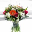 Capturing the spirit of a bright Stanmore day, this vibrant bouquet brims with extraordinary colour and freshness, as if inspired by the gardens of RAF Bentley Priory. At the centre glows a lively orange Gerbera daisy, radiating warmth and joy, surrounded by a chorus of plush crimson and magenta roses-some tightly set, others blooming with confidence-each petal inviting touch. Creamy white alstroemeria, etched with distinctive stripes, arc gently outward, intertwining with petite daisy chrysanthemums that soften the composition and add an airy lift. Honeyed hues from pale peach and yellow roses mingle above, while touches of lavender and violet peep through, adding subtle depth as if echoing Stanmore's woodland borders. Generous, glossy leaves encircle the bouquet, their sculptural beauty harmonising with slender, upright grasses that create a flowing movement. Crystal-clear water in the glass vase reflects both the pristine stems and the bouquet's fresh-picked vibrancy, promising enduring beauty for same-day delivery throughout Stanmore. Designed for moments of joy-celebrations, heartfelt thank-yous, or brightening a neighbour's day-this stunning arrangement bursts with the natural elegance and welcoming feeling that define Stanmore's local floral style.