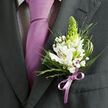 This detailed close-up shows a refined formal outfit that could be worn to a Stanmore wedding, prom at a nearby school, or a special family celebration along The Broadway. The focus rests on the left lapel of a dark charcoal grey suit, its fabric subtly textured and crisply pressed, suggesting a well-loved suit reserved for important moments. Beneath the jacket, a pale dress shirt patterned with an almost invisible dark check adds gentle visual interest, while a smooth, lavender-purple tie falls in a classic knot, catching the light with a soft satin glow. Pinned precisely to the lapel is a beautifully balanced boutonniere, the kind of piece a local florist might prepare for same-day collection before a ceremony in Canons Park. Several white alstroemeria flowers open gracefully, their petals marked with fine reddish-brown lines and small yellow throats, each bloom carefully angled to face outward. Standing tall in the centre, a conical green flower spike or bud, reminiscent of tuberose, introduces height and a fresh, lime-toned accent that plays against the coolness of the suit. Around it, clouds of delicate white gypsophila weave between the larger flowers, lending a light, airy texture, while slender grass-like fronds and broader, glossy leaves form a lush, green framework. At the base, a neatly tied satin ribbon in soft lilac encircles the stems, mirroring the lavender tie so that everything feels purposefully coordinated. The photograph conveys not only the visual beauty of the flowers, but also the quiet excitement of someone dressed and ready for a heartfelt occasion in Stanmore.