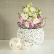 Captured in the soft, filtered daylight familiar to Stanmore's homes, this handcrafted floral arrangement radiates gentle elegance and local character. The scene centres on an airy, spherical vase-shaped from white rattan or finely painted branches-overflowing with a medley of creamy white and pale yellow lisianthus, their layered, ruffled petals inviting the touch and reminiscent of spring mornings by Stanmore Golf Course. Whispers of pale pink stock flowers add height and a sense of lush rush, while lively violet fillers-likely aster or allium-bring bursts of colour and depth, artfully bordered by fresh, dark green leaves that mimic the area's leafy paths. On the pristine, reflective white tabletop, two decorative spheres crafted from light-toned wood or rattan echo the vase's openwork texture, reinforcing an artistic and cohesive theme. Behind, a soft beige wall covering with subtle vertical patterning ensures the bouquet's pastel vibrance takes centre stage. This arrangement, imagined with the celebrations of Stanmore in mind, offers a sense of freshness, tranquility, and creative warmth-ideal for weddings or gatherings where natural form and refined craftsmanship are prized. The artistry evident in every detail showcases the care and expertise familiar to those who cherish floral delivery in Stanmore.