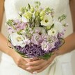 Bathed in gentle, natural light, this Stanmore bridal bouquet recalls the serene beauty of Wood Lane in early spring. It is held by hands lovingly manicured in pale blush, fingers curved protectively around the gathered stems. At the bouquet's peak, creamy white lisianthus open in ruffled, velvety waves; their soft centres echo the green freshness of a morning garden. Nestled among them are fragrant stocks in tones of delicate pink and misty lavender, lending texture and a subtle, sweet perfume to the ensemble. A dramatic band of violet allium surrounds the base, its pointed, star-like blooms creating a playful edge and celebrating the bold spirit found at a local Stanmore fête. Above, sculptural loops of deep green bear grass sweep around the floral core, giving the bouquet a sense of agility and grace. Hints of lace on the blurred wedding dress in the background suggest formality and tradition, yet nothing distracts from the arrangement's layered beauty. Designed for those who cherish both the elegance of classic wedding flowers and the freshness of contemporary Stanmore style, this bouquet is a living ode to love and artistry in the heart of North West London.