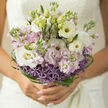 An elegant, hand-tied bouquet is held close, captured in soft, natural light that feels reminiscent of an early afternoon wandering the leafy streets of Stanmore. At the heart of the arrangement, velvety white lisianthus blooms unfurl in loose, layered cups, their petals edged with a gentle curl that catches the light. Intertwined with them, stocks in shades of pastel lilac and the faintest blush-pink create a dreamy, cloud-like fullness, their stems disappearing into a more structured base of delicate purple allium. The allium heads form starburst domes, each one composed of countless tiny florets that add a subtle sparkle and texture, anchoring the airy top of the bouquet. Around the outer edge, slender ribbons of glossy green bear grass spiral and weave, looping in graceful arcs that give the design a modern, sculptural finish. Unopened green buds nestle among the blossoms, suggesting petals still to come and moments yet to unfold. The palette is kept intentionally gentle-creamy whites, soft lilac, and fresh green-evoking the calm of a peaceful walk past Stanmore's gardens at dusk. You can almost sense the fragrance: the soft, sweet perfume of stock mingling with the delicate, leafy freshness rising from the stems. Every element speaks of careful craftsmanship, from the precise spiralling of the stems to the way each flower is placed to allow space and breath between them. It feels like a bouquet designed for the first quiet minutes of a wedding day, perhaps waiting in a Stanmore townhouse or near the station, embodying tenderness, optimism, and an understated, enduring romance.