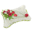 A carefully made sympathy cushion, presented as a soft rectangular pillow entirely cloaked in hundreds of small, pristine white chrysanthemum blooms, fills the frame with a sense of hush and peace. The densely packed heads create a velvety, cloud-like surface that invites the hand to imagine a gentle texture; under soft daylight the white petals read pure and unblemished, catching faint shadow in the hollows between blooms. Around the edge a narrow band of pale green satin ribbon peeks out, giving a subtle, fresh definition to the pillow's contour and finishing the piece with an understated trim. Anchoring the design at the upper left is a rich spray of deep red roses and ruffled burgundy carnations, their velvety petals and layered forms providing a warm, reverent contrast to the white field. Interspersed among them are small clusters of creamy white accent flowers-freesias or spray chrysanthemums-that add delicate highlights and a whisper of perfume. Lush green foliage, including feathery fern fronds and broader leaves, lifts the arrangement and frames the blooms with verdant depth. From that principal cluster, elegant arcs of bright green bear-grass or similar slender foliage sweep diagonally toward the lower right, creating a graceful line and suggesting movement across the pillow. The sweep culminates in a single, perfectly shaped red rose nestled on the lower corner, as if carried there by those green strokes-a poignant symbol of enduring love. Crafted with quiet care, this traditional funeral pillow would be fitting for services held in Stanmore's churches or at nearby memorials, offering comfort, respect and calm remembrance to grieving families.