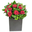 In this elegantly modern composition, deep crimson roses command the centre with a composed, velvety intensity. Each bloom is full and tightly spiralled, petals curling inward in soft, generous layers that catch the light and read as almost tactile velvet. Around them, broad glossy leaves have been looped and folded with a florist's restraint - sculptural ribbons of vibrant green that move in overlapping waves, adding rhythm and depth without stealing from the roses' drama. The whole sits in a matte black square vessel, its geometric lines providing a confident, grounded contrast to the organic curves above. Studio lighting here is clean and steady, picking out the sheen on each leaf and the subtle highlights on the red petals so the crimson reads warm and saturated, like a pause in a busy day. There's a faint, fresh green note that lifts the classic rose perfume into something lighter - a breath of garden-fresh foliage that tempers the sweetness and makes the bouquet feel poised rather than heavy. As a florist who often walks Stanmore High Street and knows the quiet mornings by Canons Park, I imagine this arrangement placed on a small café table or a city apartment console: modern, considered, and quietly affectionate. It's the kind of piece offered for anniversaries, intimate congratulations, or to brighten an office trolley - a visual and fragrant reminder that simple, expertly handled materials can communicate warmth and intention without fuss. The overall effect is contemporary romance: bold in colour, refined in structure, and deeply rooted in thoughtful craft.