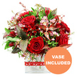 A beautifully composed bouquet in passionate reds and complementary greens stands out clearly against a white background, conveying the feel of a romantic gift poised for delivery anywhere in Stanmore. At the heart of the design, velvety red roses are arranged in a cluster, each bloom telling its own story-some buds still tightly furled, others gently opening to reveal layers of soft, overlapping petals. Around these central roses, smaller deep crimson flowers, reminiscent of alstroemeria or similar multi-petalled varieties, are artfully tucked, creating a rich tapestry of shapes and tones. Threading through the arrangement are fine sprays of tiny waxflowers or limonium in pale pink and creamy white, their numerous small, star-like blossoms softening the overall look and adding delicate detail up close. A generous halo of greenery frames the flowers, with glossy dark green leaves, lighter sprigs, and distinctive burgundy and reddish-brown foliage with subtle veining lending depth, contrast, and a sense of natural abundance. The bouquet is crafted into a rounded, dome-shaped composition that feels full and balanced from every angle, as if designed to sit at the centre of a dining table or sideboard. The stems are placed carefully in a sleek, clear glass vase with a modern rectangular base, their clean lines visible through the water and reinforcing the contemporary style. A bright red ribbon, printed with small white heart outlines, wraps around the vase and is tied neatly, underscoring the romantic intention-ideal for anniversaries, Valentine's Day, or a heartfelt surprise for someone living near Canons Park or along the quiet residential streets of Stanmore. In the lower right corner of the image, a bold circular orange overlay displays the words VASE INCLUDED in crisp white uppercase letters, clearly indicating this is a complete, ready-to-display floral gift.