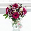 A vibrant mixed bouquet of fresh flowers stands proudly in a clear, textured glass vase, placed against a softly glowing white background that suggests daylight streaming through a Stanmore window. At the heart of the arrangement are large, velvety gerbera daisies in deep fuchsia and magenta tones, their dark centres drawing the eye and giving the bouquet a joyful, confident energy. Around them cluster bicolour chrysanthemums, each petal tipped in rich purple that fades delicately to white, framing a bright yellow centre and adding a sense of playful movement. Creamy white spray roses, some fully open and others still in tight bud, bring a gentler, romantic note, their petals appearing almost satin-smooth. Wisps of tiny white gypsophila are threaded throughout, creating a light, cloudlike texture that softens the bold colour palette. Lush green foliage cradles the blooms and spills neatly over the rim of the vase, grounding the design in nature. The squat jar-style vase is tied at the neck with a simple rustic twine bow, from which hangs a small, white rectangular gift tag printed with a soft pink rose motif and graceful green lettering, suggesting a thoughtful message from the sender. The bright, diffused lighting enhances every detail-the glass catches subtle reflections, the greens appear freshly cut, and the petals look dewy and new, as though the bouquet has just arrived from a local florist off Stanmore Broadway. Overall, the arrangement radiates warmth and celebration, perfect for a birthday surprise, a cheerful thank-you, or simply brightening a home near Canons Park or Stanmore Hill with a burst of colour and life.