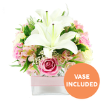 A compact yet richly detailed bouquet of white lilies and pink flowers is shown against a simple white background, its colours and textures clear and inviting, as if freshly arranged at Flower Delivery Stanmore before heading out on a local delivery. Three white Oriental lilies take pride of place in the centre, their generous petals opening upward and outward, revealing soft yellow-toned stamens tipped with darker hues that draw the eye to their fragrant hearts. Around them, numerous pink roses cluster closely; some are tight buds with neatly wrapped petals, while others are open and ruffled, revealing soft, velvety layers that speak of affection and thoughtful care. Scattered throughout, peach-pink alstroemeria blooms add gentle warmth, their slender petals marked with fine speckles and stripes that lend subtle complexity. Tiny white gypsophila blossoms float among the larger flowers like small white stars, lightening the arrangement and giving it a dreamy, cloud-like aura. A selection of green foliage-broad, smooth leaves and finer, more delicate stems-frames the bouquet and adds a sense of natural freshness, evoking the greenery found around Stanmore's quiet residential streets and local parks. The flowers are arranged in a clear, square glass vase that exposes the trimmed stems in the water, reflecting a clean, contemporary style. Around the middle of the vase, a pale pink satin ribbon is tied in a simple horizontal band, offering a soft, romantic accent. In the lower right part of the image, a circular orange emblem bears the words VASE INCLUDED in bold white capital letters, clearly signalling that the glass container comes with the arrangement. The overall effect is one of gentle joy and refined elegance, ideal as a ready-to-display gift for Mother's Day, anniversaries, or a comforting gesture delivered anywhere in Stanmore.
